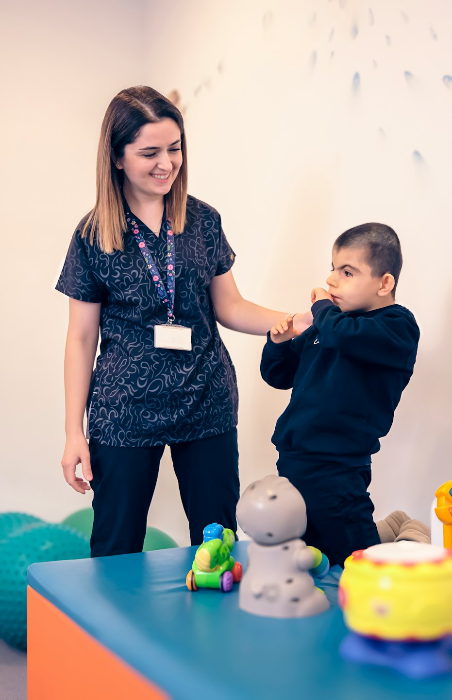 Woman guides a young boy's arm in a therapy room.